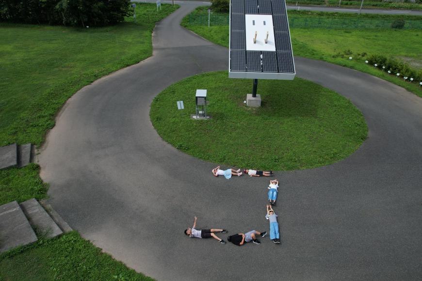 地面に寝ころびカタカナのコの字を作っている子供たち6人を上空からドローンが撮影した写真