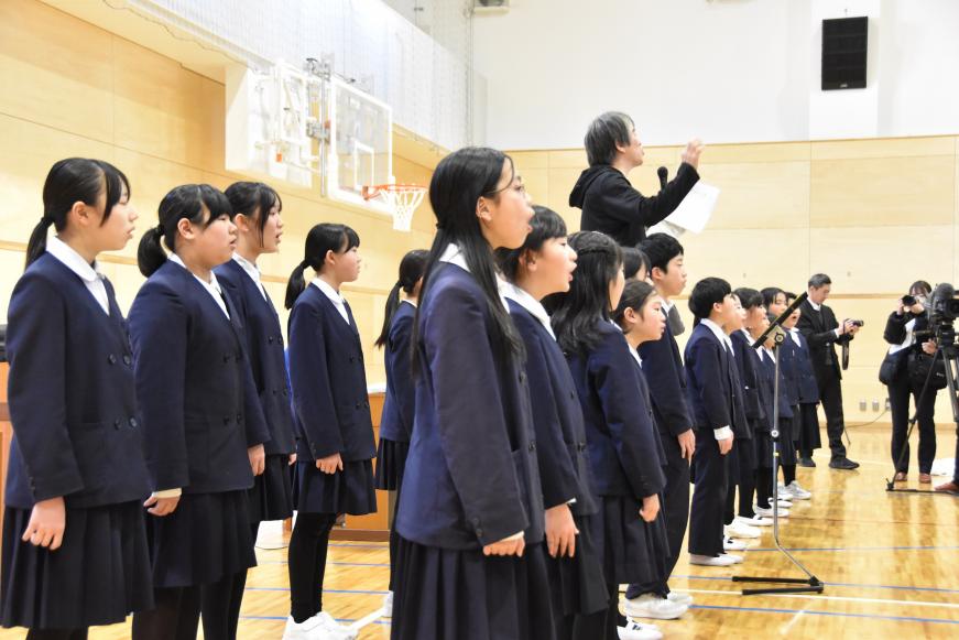 合唱部児童が着席する生徒達の前で先生の指揮に合わせて合唱している写真