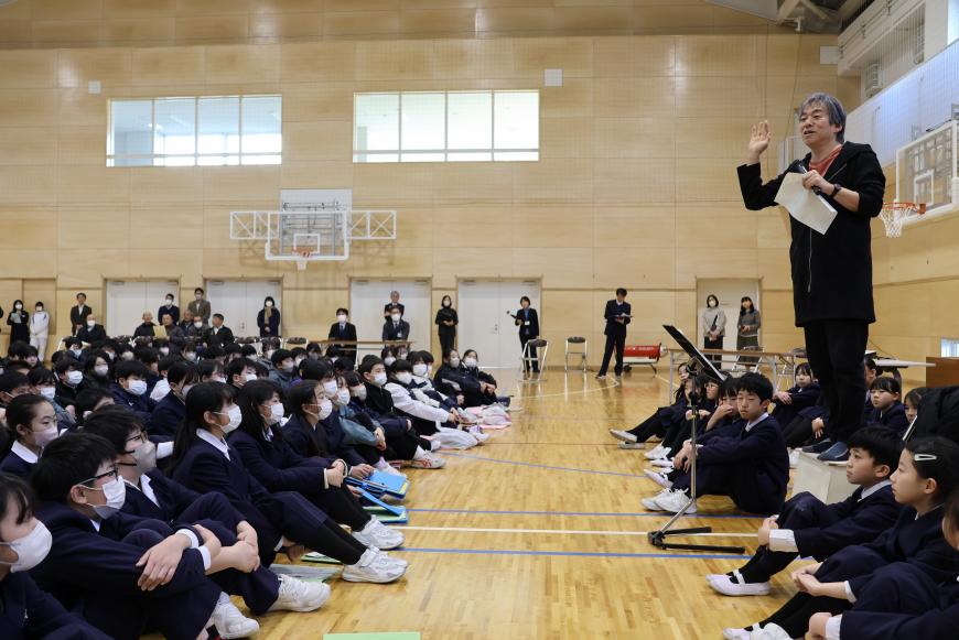 男性の先生が立って話をするのを着席する生徒達が聞いている写真