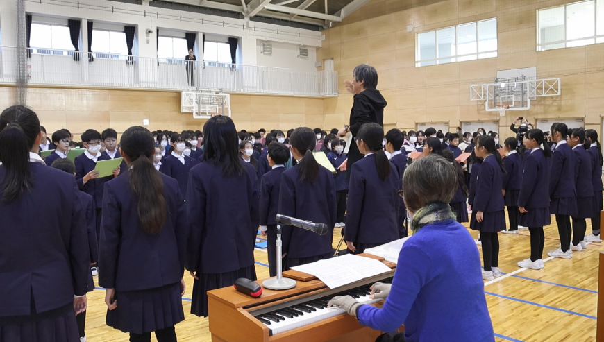 合唱部児童達と他の生徒達が向かい合わせに整列し男性の先生の指導の元、合唱している様子の写真
