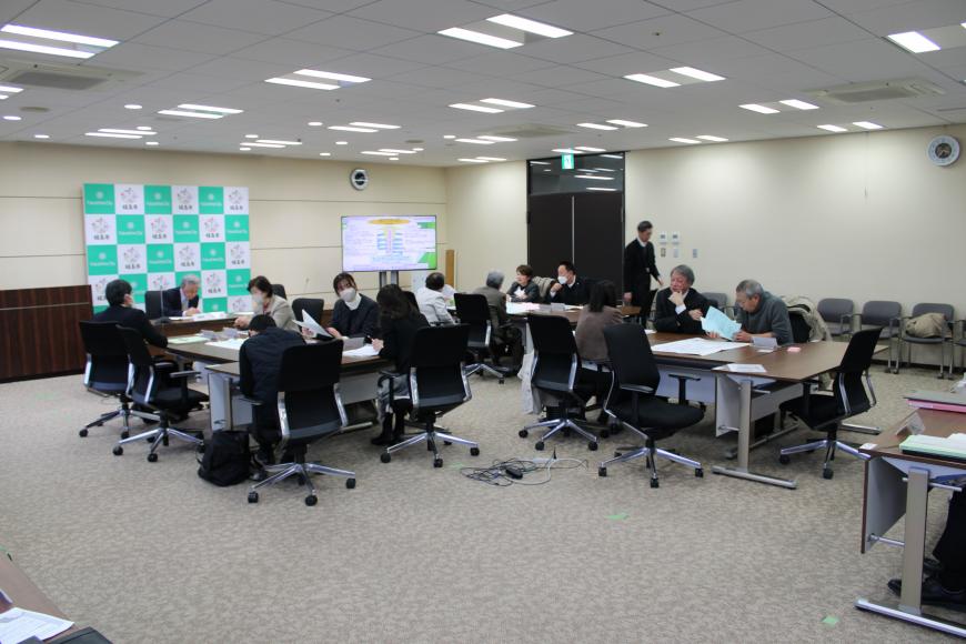 参加者達がグループ後ごとに着席し、前のスクリーンの資料を元に会議が行われている写真