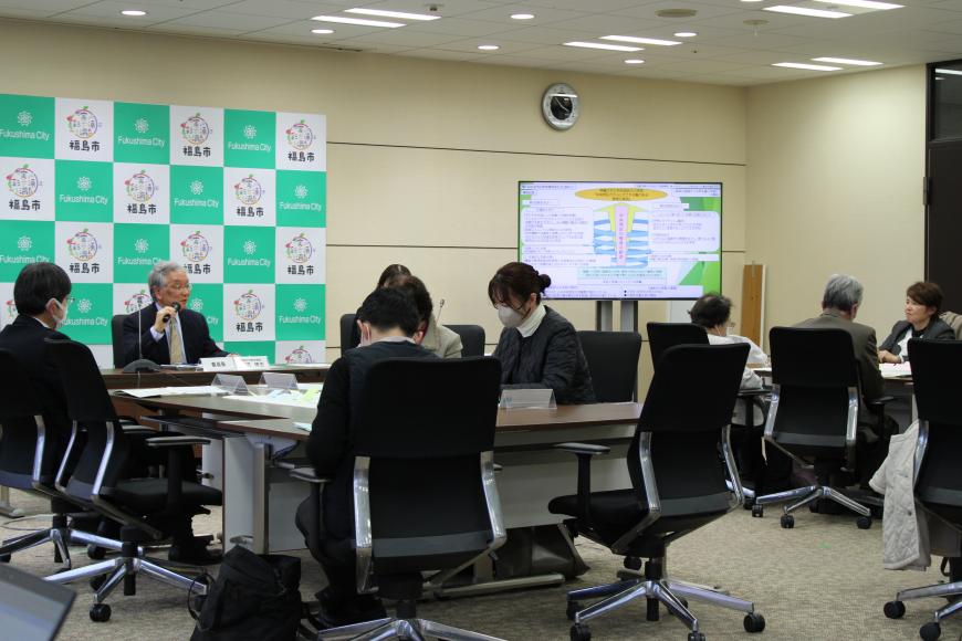 参加者達がグループ後ごとに着席し、前の男性がマイクで話をしており、スクリーンの資料を元に会議が行われている写真