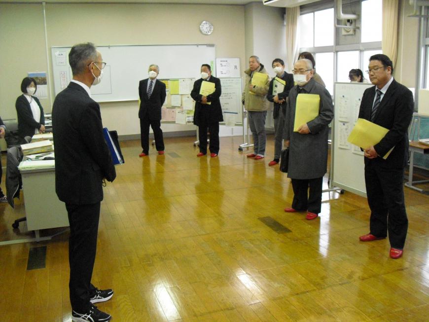 前方にスーツを着た男性が立って話しているのを、資料を持った関係者達が聞いている様子の写真