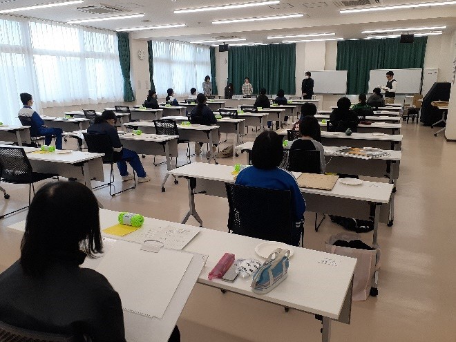 テーブルと椅子が並べられた室内で、前方で男性が立って話しているのを参加者がそれぞれの席に着席し聞いている写真