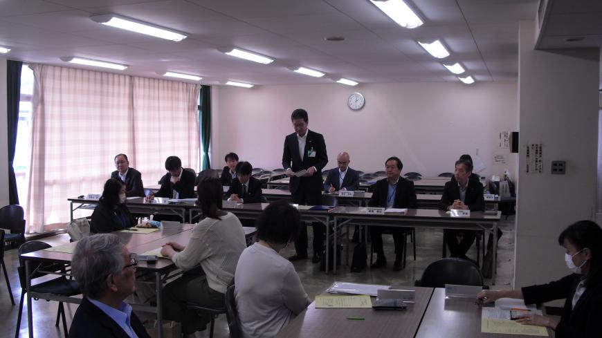 前方で男性が立って話をしているのを、グループごとに設置された長机と椅子に着席した参加者達が聞いている様子の写真