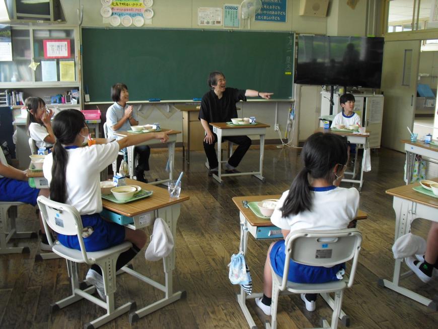 机で配膳された給食を食べており、女の子と男性教師が何かを指差して笑っている様子の写真
