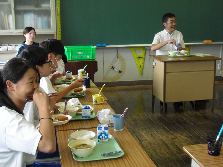 机に配膳された給食を食べている生徒たちと先生の写真