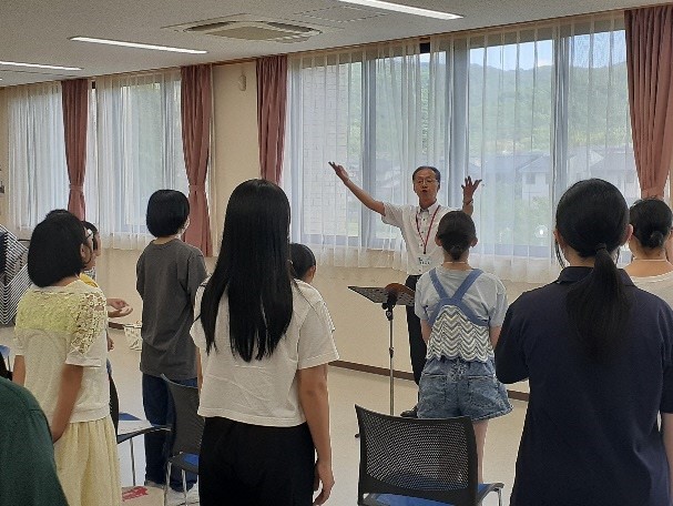両手を広げた男性指導者と向かい合わせに並んで練習をする参加者達を後ろから撮影した写真