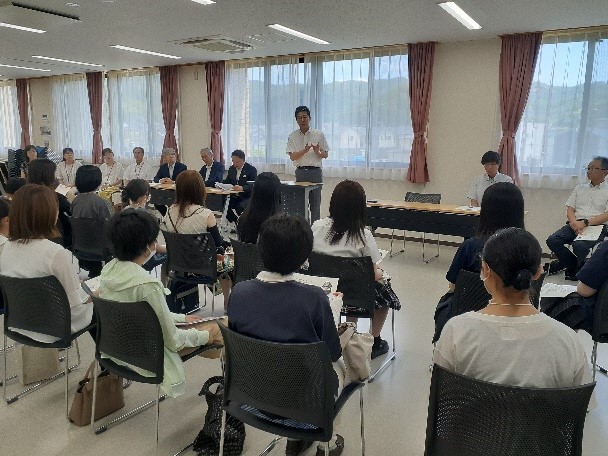 男性指導者が立って説明し、横に関係者が座り、参加者達が椅子に座り話を聞いている写真
