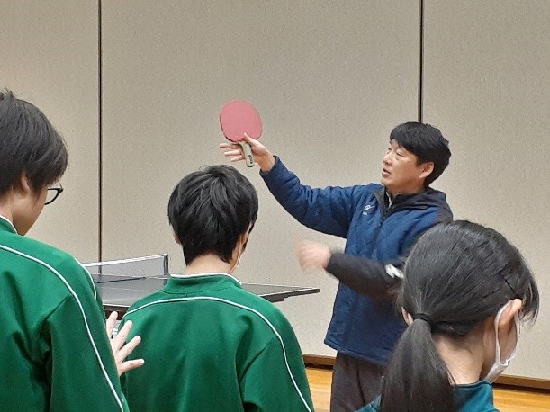 指導者がラケットを持って説明するのを聞いている参加者達の写真