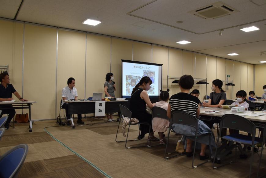 スクリーンに映し出された資料を元に講師の女性が立って話をするのを、着席した参加者が聞いている写真