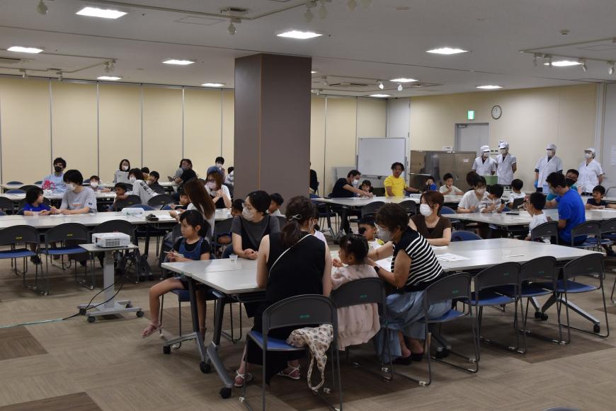 白い帽子と白衣を着た職員達が4人後ろに立っており、食育の話を聞いている参加者の写真