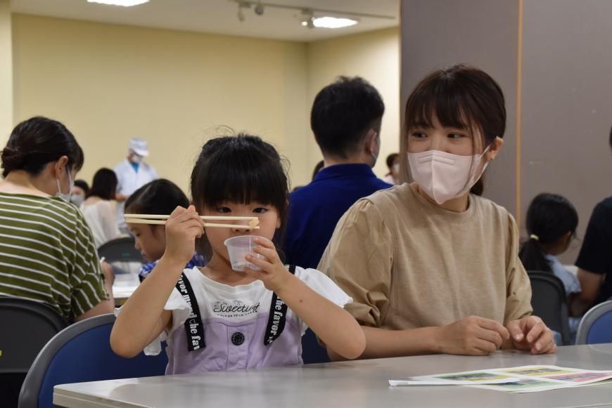 女の子が透明の容器を左手で持ち、右手で箸を使い粒をつかんでいる様子を見ているお母さんの写真