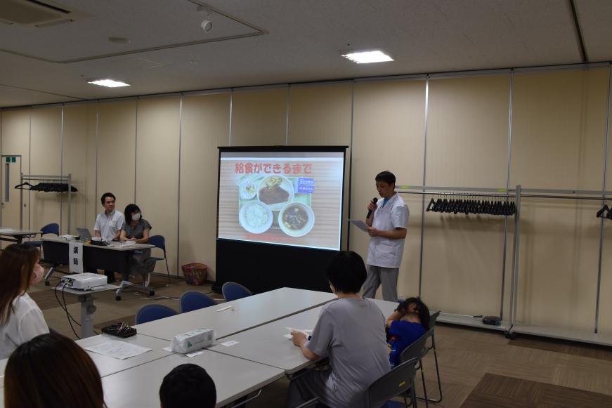 スクリーンに「給食ができるまで」の資料が写し出され、関係者が横に座り、マイクを持った男性が説明をしているのを参加者が聞いている様子の写真