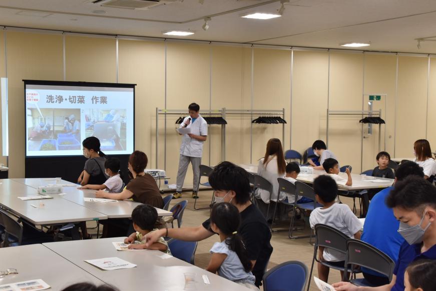 スクリーンに「洗浄・切菜・作業」の資料が写し出され、マイクを持った男性が説明をしているのを参加者が聞いている様子の写真