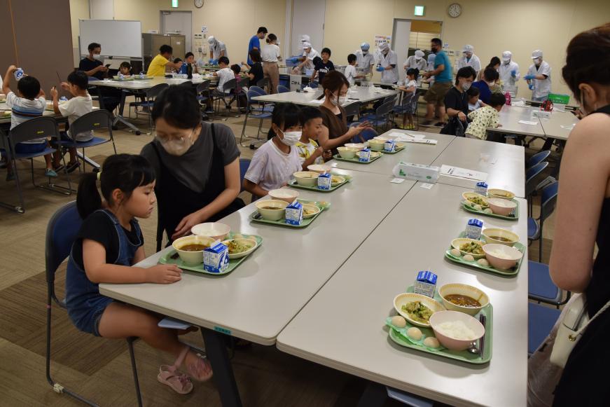 テーブルに配膳された食事が置かれ参加者達が着席し、各テーブルに分かれて試食の準備をする様子の写真