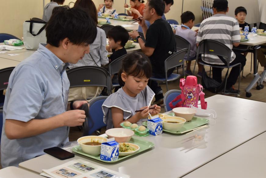 テーブルに食事が置かれ、女の子が箸を両手で持っているのを見ている親子の写真