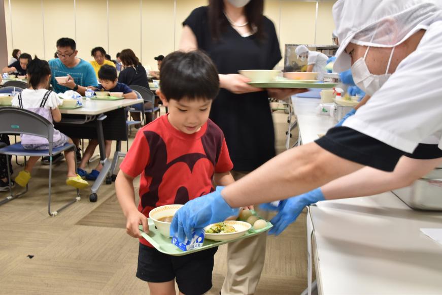 赤いTシャツを着た男の子が持つトレイに職員の方が牛乳をのせている写真