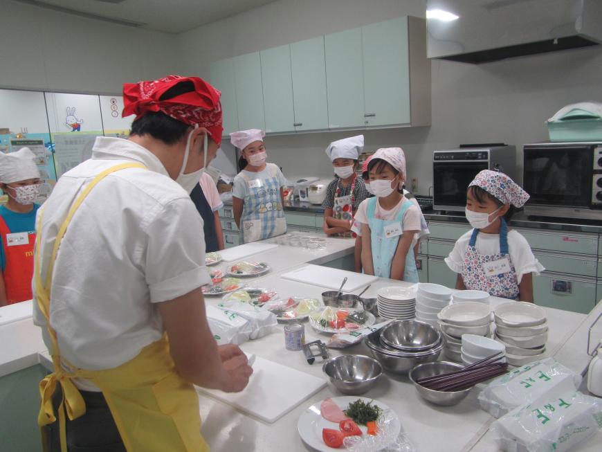 赤の三角巾とエプロンを身につけた調理職員が野菜を切る様子を真剣な表情で見ている子供たちの写真