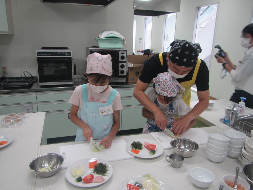 調理職員と一緒に野菜を切る女の子、野菜に手を添えて包丁で野菜を切る女の子の写真
