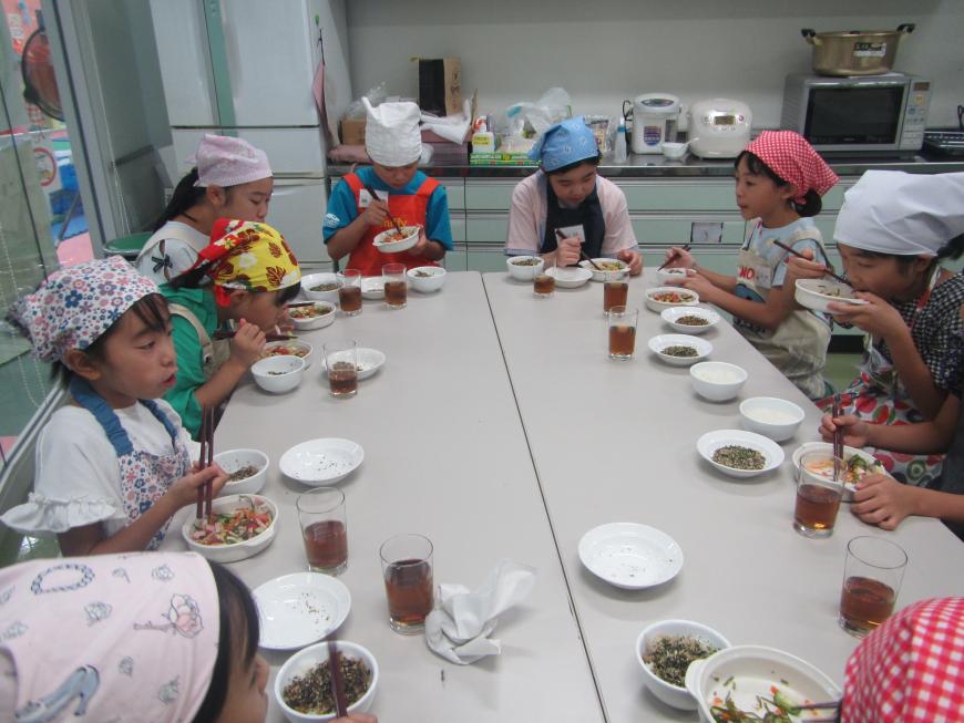 三角巾とエプロンを身につけた子供達が着席し作った料理を試食している写真