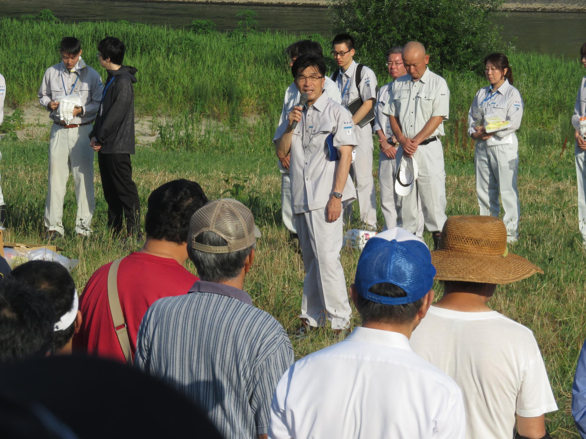 河川敷でグレーの作業服を着た関係者たちの前に立った、作業服姿の市長がマイクを持ち参加者たちへ話をしている様子の写真