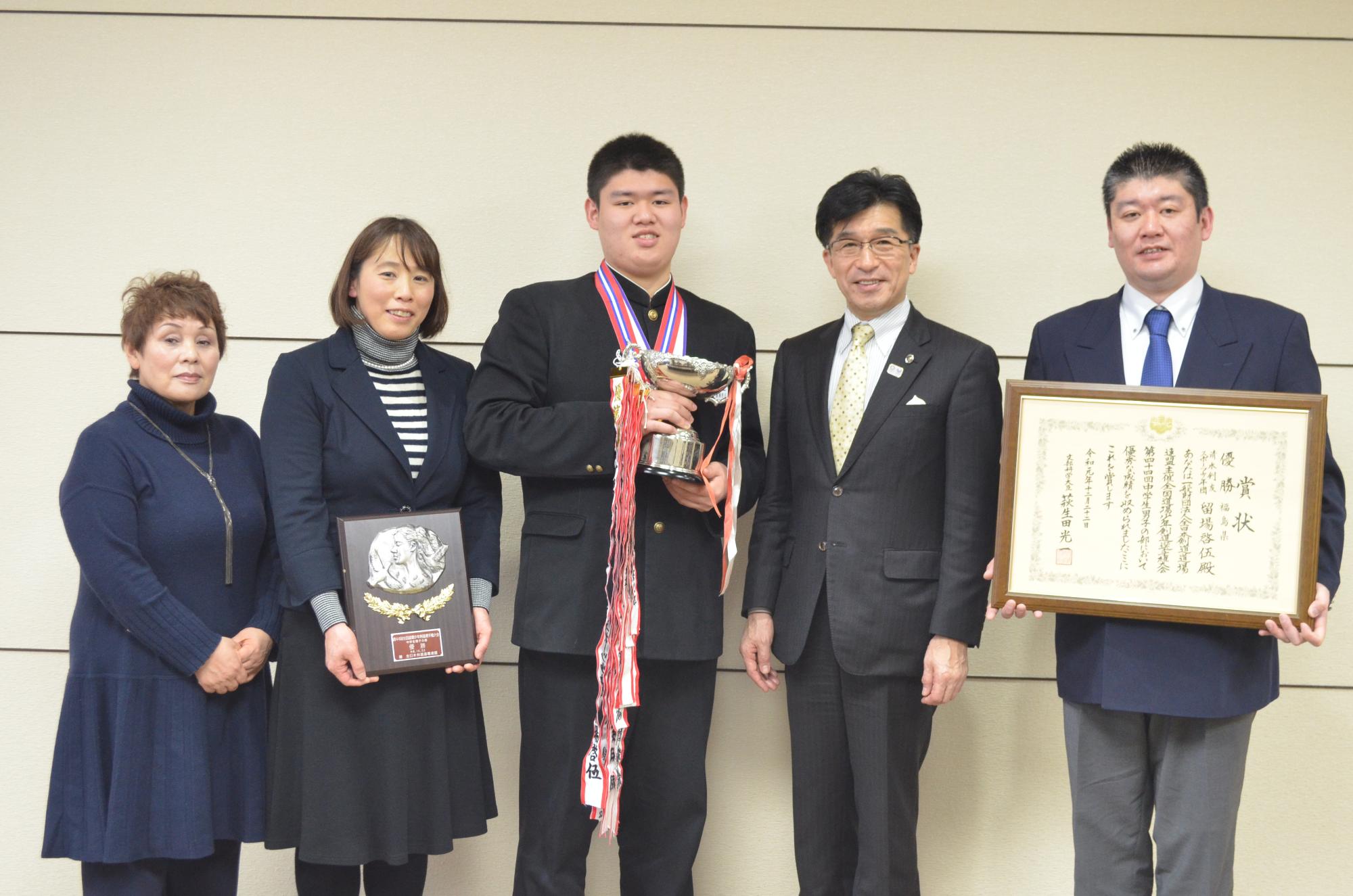 トロフィーを持った留場選手の隣に女性2名が立ち、うち1名は表彰楯を手に持っており、反対の隣には、市長と賞状を持った男性が立ち、横並びで記念撮影をしている写真