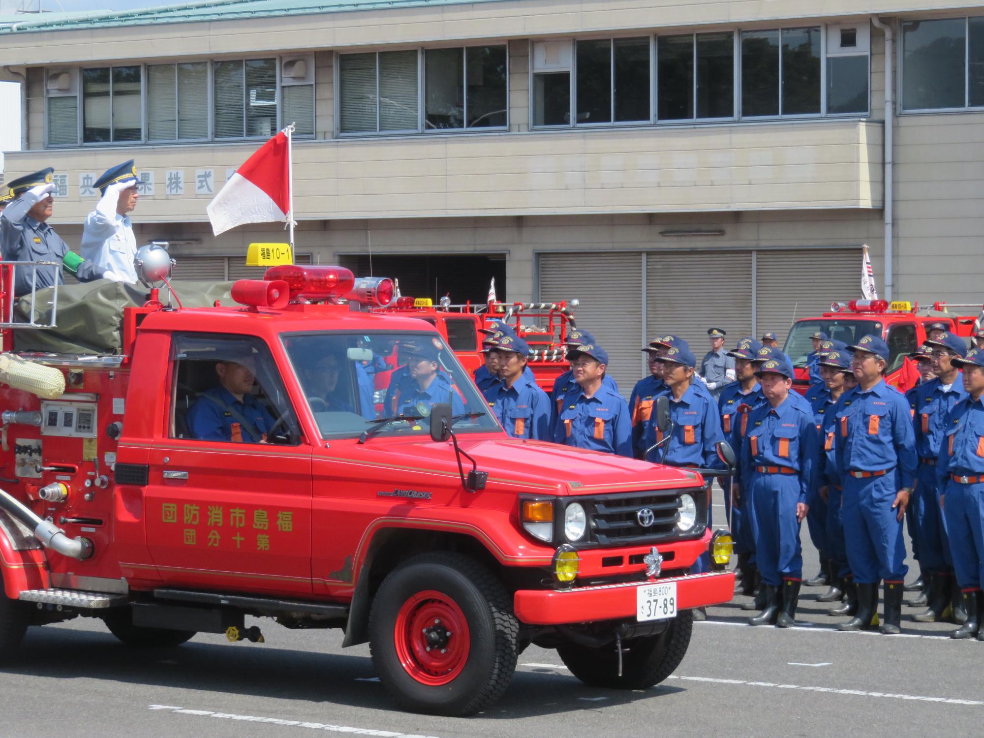 白い壁の建物の前に消防車両と青い制服の消防団員が整列し、その前を敬礼をする団員を乗せた消防車両が走っている様子の写真