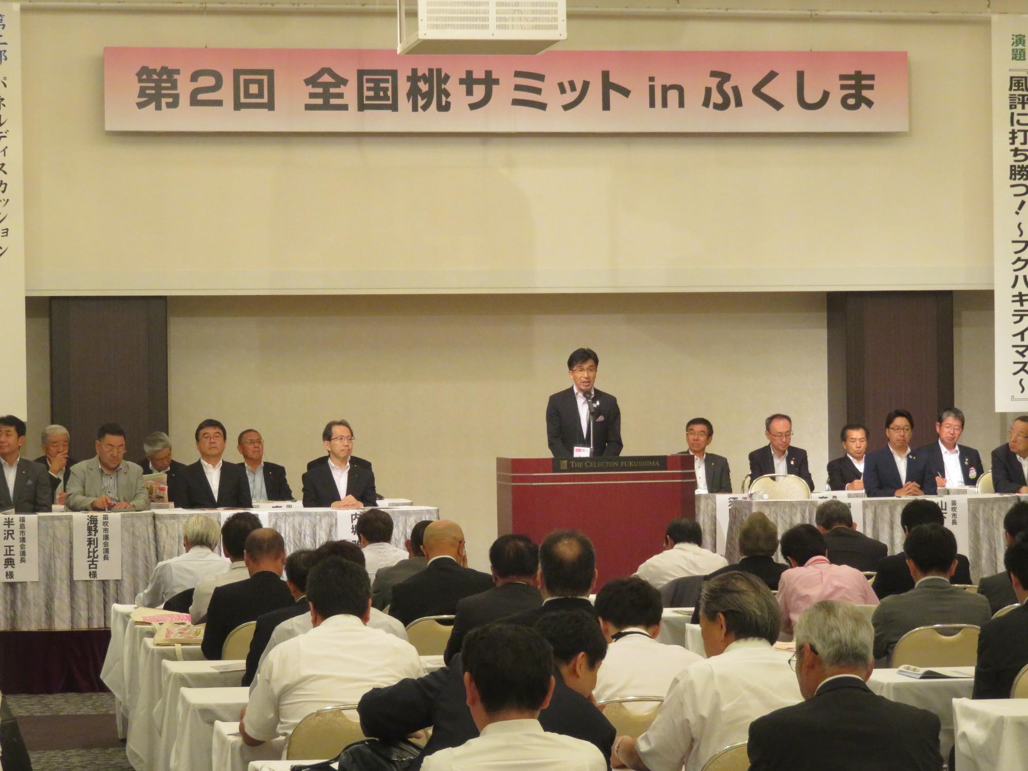 「第2回全国桃サミットinふくしま」の看板が掲げられた会場で、左右の関係者席の間の演台に立ち前に座った出席者に向かって話をする市長の写真