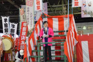 階段や壁面に紅白幕が張られており、はっぴ姿の市長がマイクスタンドの前に立ち、話をしている写真