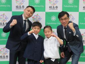 バックパネルの前で、木村さんと市長、熊坂さんと菊田さんがガッツポーズをして横一列に並んでいる写真