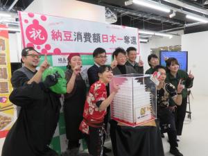 「納豆消費日本一奪還」と書かれたバックパネルの前で、透明のケースに入った白い模型の様な物を囲んで市長や9名の関係者が人差し指で1のポーズをし並んでいる写真
