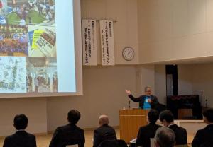 会場前方に設置されたスクリーンに資料が映し出され、右側の檀上で右手を広げながら講演を行っている岩本唯史さんの写真