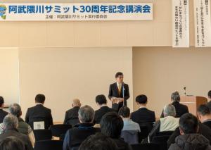 「阿武隈川サミット30周年記念講演会」と書かれた横断幕が掲げられた会場の前方に立つ市長が挨拶を行っている写真