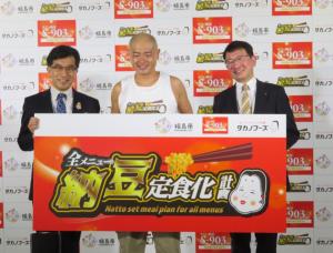 「全メニュー納豆定食化計画」と書かれたボードの後ろにあばれる君を挟んで左に市長と右に関係者の男性が横一列に並んでいる写真