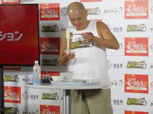 バックパネルの前でペットボトルの水や食器が置かれた白い丸テーブルの前に立つあばれる君がかき混ぜた納豆を伸ばしている写真