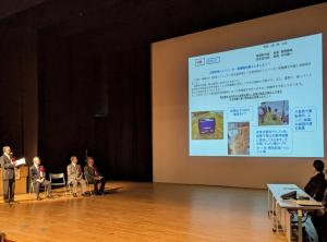 大きなスクリーンに資料が映し出されたステージ左右に関係者の方々が座り、左端に立っている男性が話をしている様子の写真
