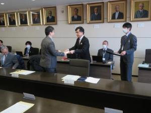 横長のテーブルが配置された室内で、グレーのスーツを着た男性と市長が向かい合って立っている場面を写した写真。