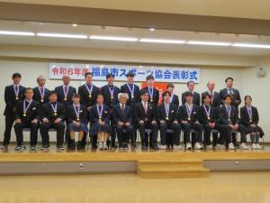 「令和6年度福島市スポーツ協会表彰式」と書かれた横断幕が掲げられた小さなステージ上の椅子に市長を中央に10人、後列に11人並んでいる記念写真