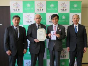 バックパネルを背に、目録を手にした市長と石本会長が中央に立ち、両側に関係者の男性が横一列に並んで撮影された記念写真