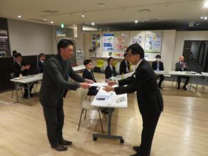 二列に並んだ長机の席に塾生が座り、塾生代表の男性と市長が向かい合あって立っている写真