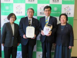熨斗がかけられたお米の袋を持った市長と、日本敬神崇祖自修団の関係者3名が、バックパネルを背に横一列に並んで記念撮影をしている写真