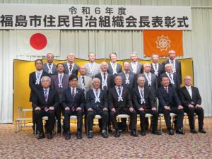 「令和6年度福島市住民自治組織会長表彰式」と書かれた横断幕と国旗と市旗が掲げられたステージ上に金屏風が設置され、前列の椅子に7人座り、2列目と3列目に7人ずつ立ち並んでいる記念写真