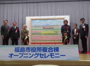 「福島市役所複合棟オープニングセレモニー」と書かれた看板が設置されたステージ上に大きな絵画が設置され、左右に胸章リボンをつけた2人の園児や市長、5名の関係者が並んでいる写真