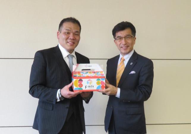 壁の前で市長と玉ノ井親方が一緒に紙製のボックスを持っている写真
