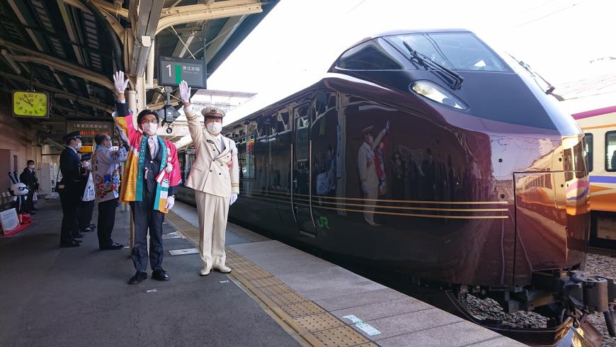 ホームに停車しているワイン色の列車の先頭車両の横で白の制服姿の駅員と法被を着た市長が並んで右手を挙げている写真