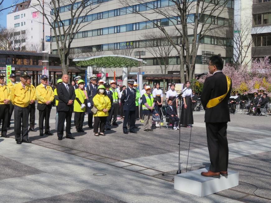 街頭で黄色の襷をかけた市長が黄色のジャンバー姿、警備員姿、反射材のベストを着た方々が整列している前で話をしている写真
