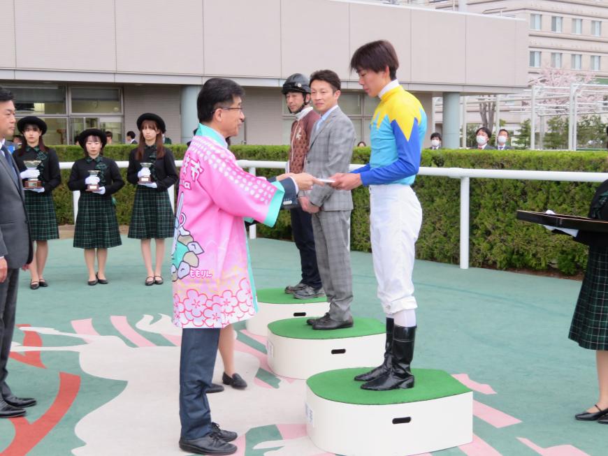 関係者の方が見ている中、法被を着た市長が台に乗った青色の服を着た騎手に目録を手渡している写真