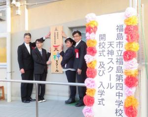 建物入口に設置された看板を、市長を含む4名で持ちカメラを向いている記念写真