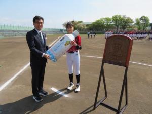 グランドで市長がユニフォーム姿の女子選手に巨大ペットボトルを渡している写真